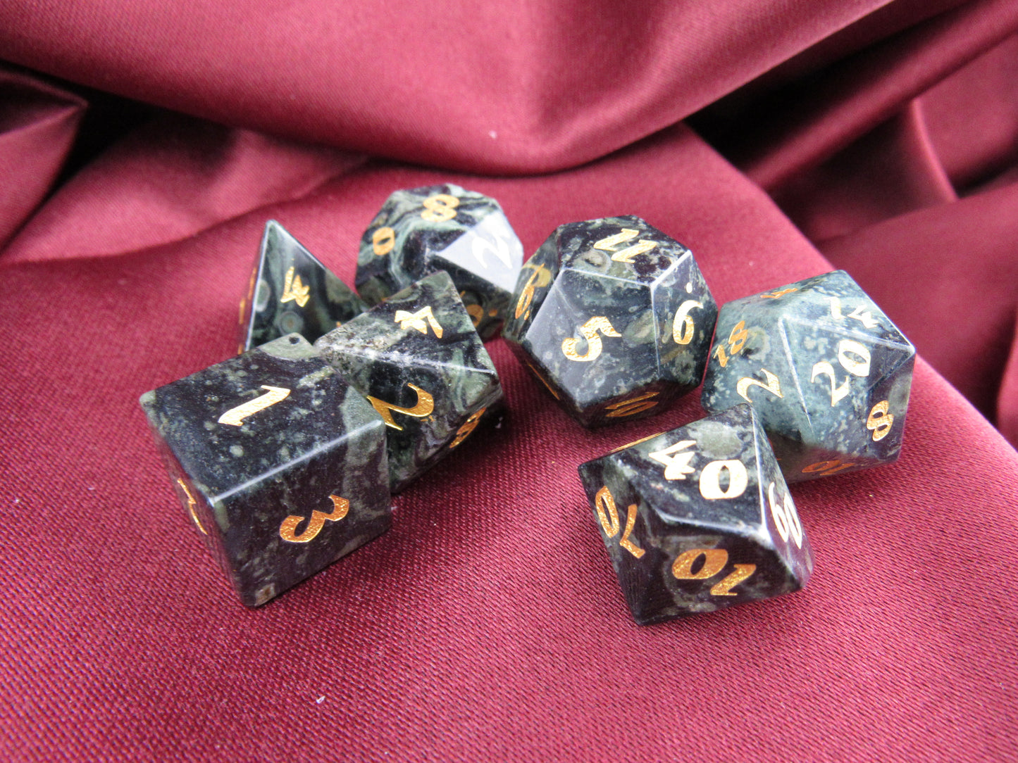 A set of seven polyhedral gaming dice with a dark gray and black mottled stone appearance and gold-colored numbers, arranged on a draped burgundy fabric background.