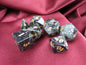 A set of seven polyhedral gaming dice with a dark gray and black mottled stone appearance and gold-colored numbers, arranged on a draped burgundy fabric background.