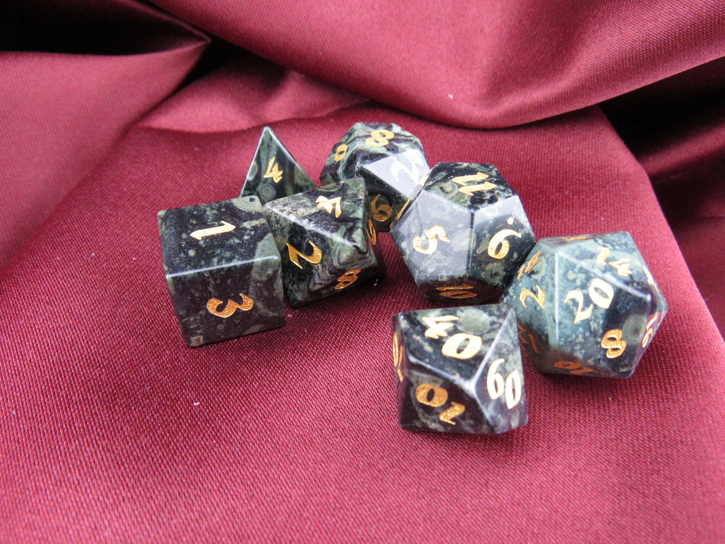 A set of seven black polyhedral gaming dice with gold numbers, arranged on a draped dark red fabric background.