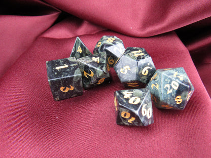 A set of seven black polyhedral gaming dice with gold numbers, arranged on a draped dark red fabric background.