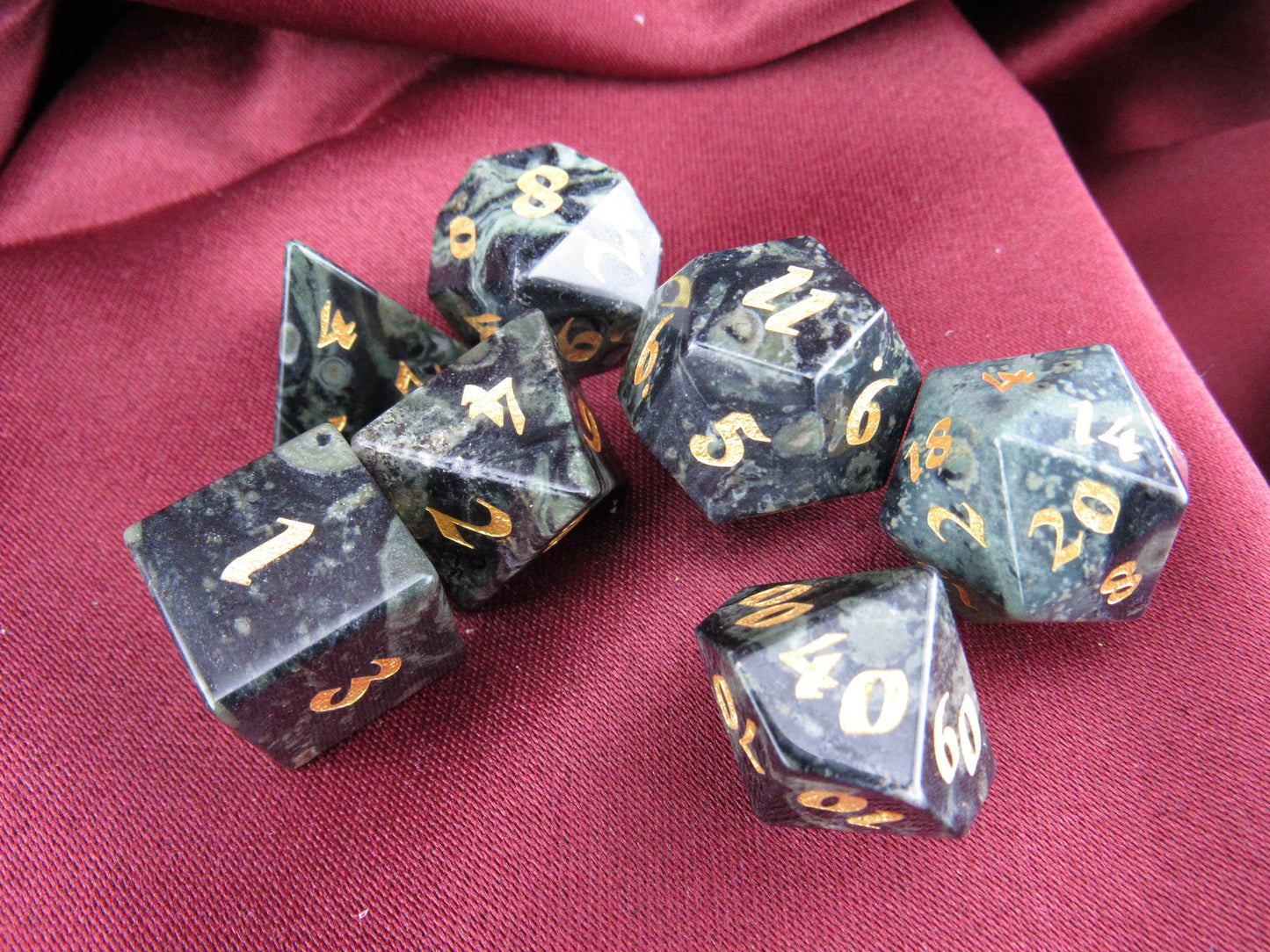 A set of seven black polyhedral gaming dice with gold numbers, arranged on a draped dark red fabric background.