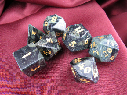 A set of seven black polyhedral gaming dice with gold numbers, arranged on a draped dark red fabric background.